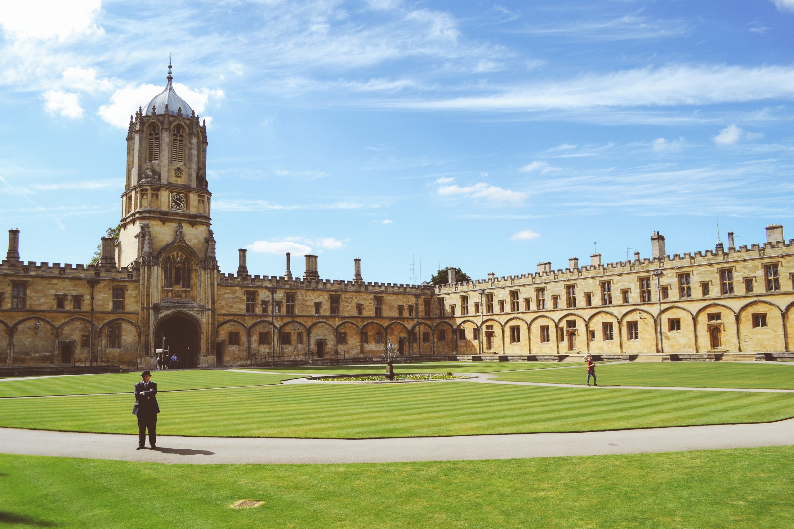 Tom Tower and the honey-coloured stone buildings of Tom Quad at Christ Church, Oxford, around a wide green lawn under a blue sky
